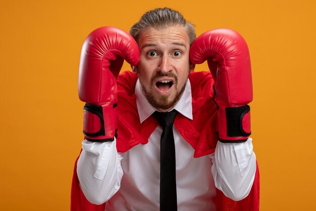 Chico joven superhéroe asustado con corbata y guantes de boxeo poniendo las manos en la cabeza aislada sobre fondo naranja