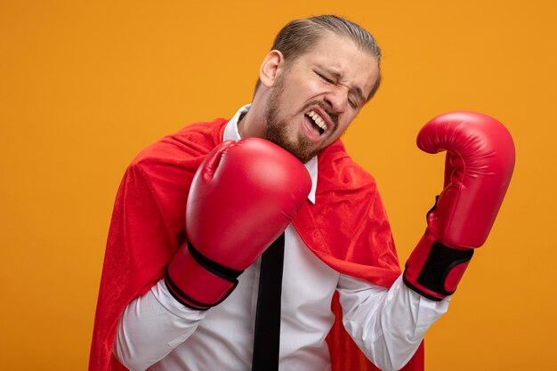 Chico joven superhéroe cansado con corbata y guantes de boxeo aislados sobre fondo naranja