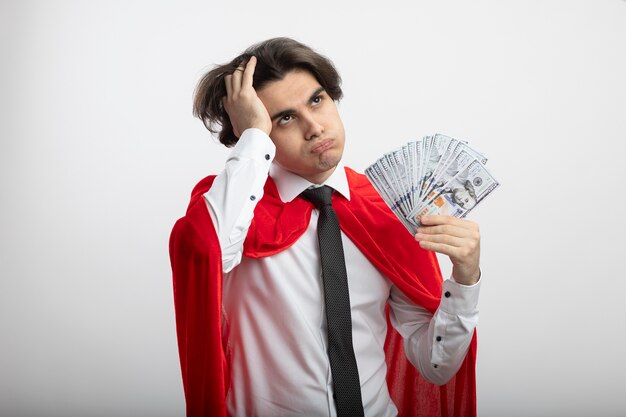 Foto gratuita chico joven superhéroe cansado mirando a un lado con corbata sosteniendo dinero en efectivo y poniendo la mano en la cabeza aislada en blanco