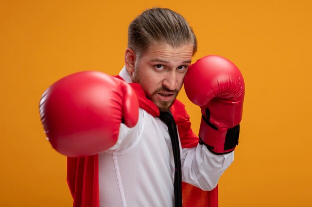 Chico joven superhéroe confiado con corbata y guantes de boxeo de pie en pose de lucha aislado en naranja