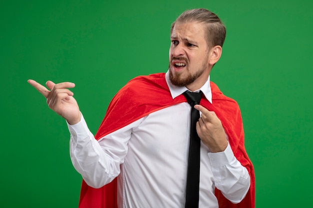 Foto gratuita chico joven superhéroe confundido con corbata mirando al lado y puntos al lado aislado sobre fondo verde