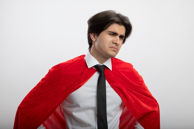 Chico joven superhéroe confundido mirando a la cámara con corbata aislado sobre fondo blanco.