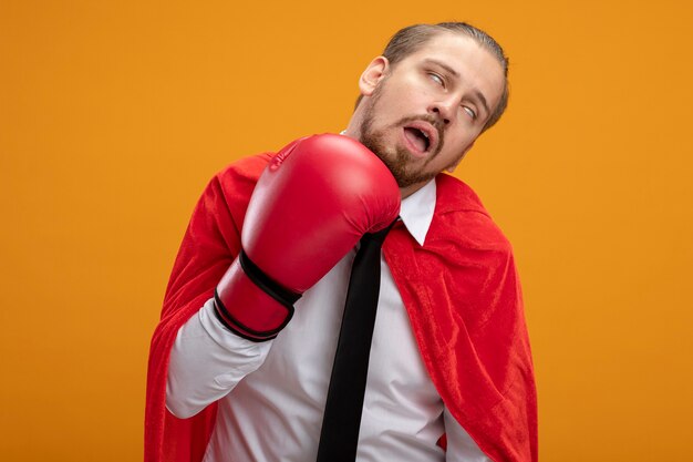 Chico joven superhéroe con corbata y guantes de boxeo golpeándose a sí mismo aislado en naranja