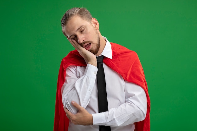 Chico joven superhéroe con corbata con los ojos cerrados mostrando gesto de sueño aislado en verde