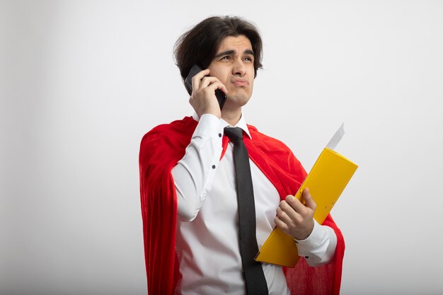 Chico joven superhéroe descontento mirando al lado con corbata sosteniendo la carpeta habla por teléfono aislado en blanco