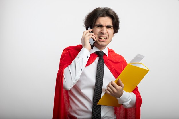 Chico joven superhéroe disgustado con corbata sosteniendo la carpeta y habla por teléfono aislado en blanco