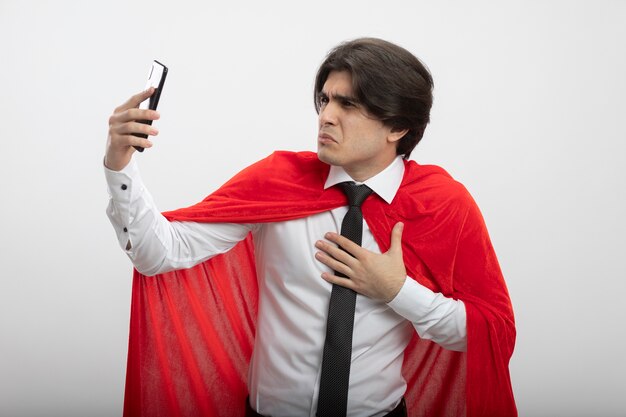 Chico joven superhéroe disgustado con corbata sosteniendo y mirando el teléfono poniendo la mano en el pecho aislado sobre fondo blanco.