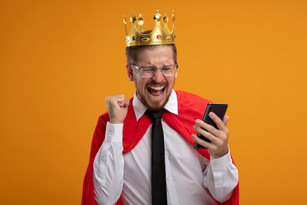 Foto gratuita chico joven superhéroe emocionado con corbata y corona sosteniendo y mirando el teléfono mostrando sí gesto aislado sobre fondo naranja