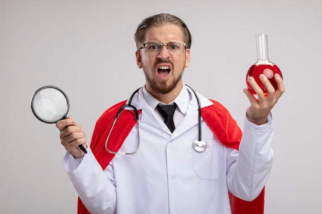 Chico joven superhéroe enojado con bata médica con estetoscopio y gafas con lupa con botella de vidrio de química llena de líquido rojo aislado sobre fondo blanco.