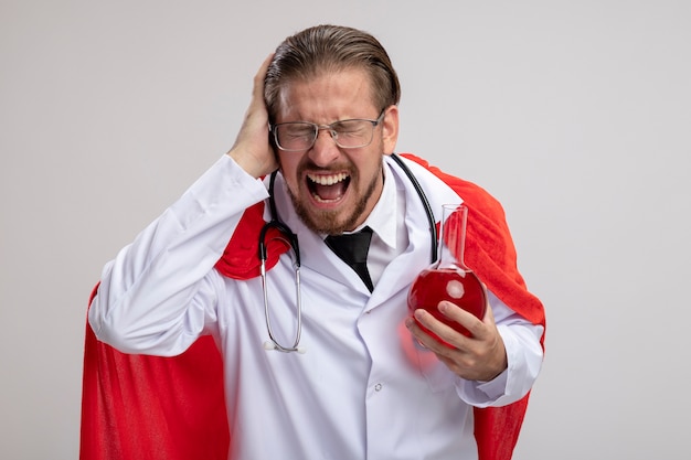 Chico joven superhéroe enojado con bata médica con estetoscopio y gafas sosteniendo una botella de vidrio de química llena de líquido rojo poniendo la mano en la cabeza aislada sobre fondo blanco