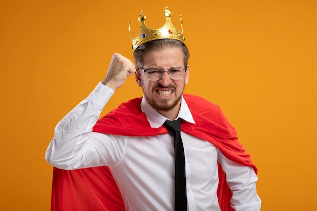 Foto gratuita chico joven superhéroe enojado con corbata y corona con gafas levantando el puño aislado en naranja