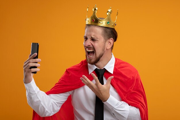 Chico joven superhéroe enojado con corbata y corona sosteniendo y mirando el teléfono aislado en naranja