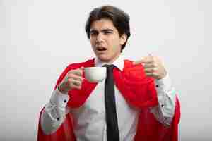 Foto gratuita chico joven superhéroe impresionado con corbata sosteniendo y puntos en la taza de café aislado sobre fondo blanco.