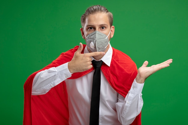 Chico joven superhéroe con máscara médica y corbata fingiendo sostener y apunta a algo aislado en verde