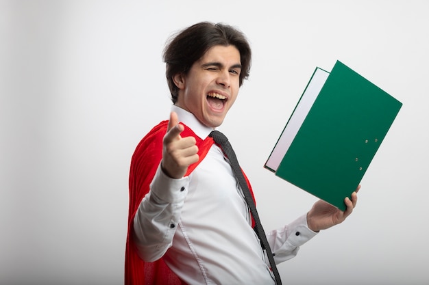 Chico joven superhéroe parpadeó alegre con corbata sosteniendo el portapapeles y mostrándole gesto aislado en blanco