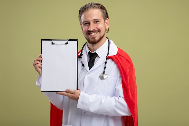 Chico joven superhéroe sonriente con estetoscopio con bata médica sosteniendo el portapapeles aislado sobre fondo verde oliva