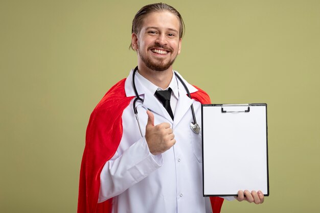 Chico joven superhéroe sonriente con estetoscopio con bata médica sosteniendo el portapapeles mostrando el pulgar hacia arriba aislado sobre fondo verde oliva