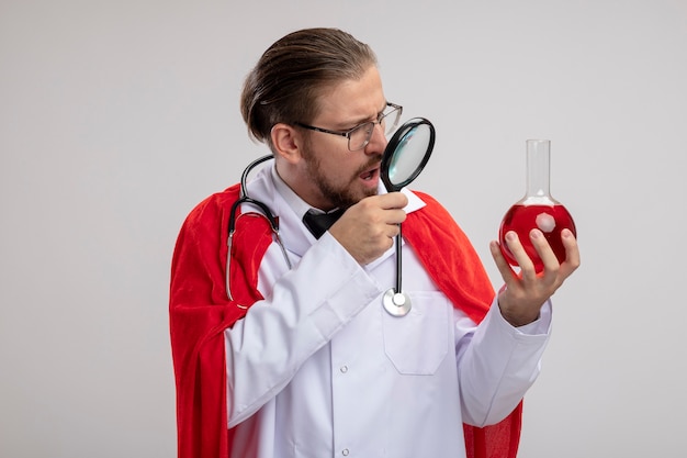 Foto gratuita chico joven superhéroe sorprendido con bata médica con estetoscopio y gafas sosteniendo y mirando la botella de vidrio de química llena de líquido rojo con lupa aislada sobre fondo blanco