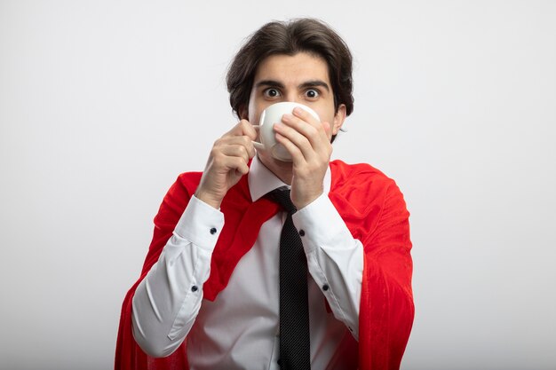 Chico joven superhéroe sorprendido mirando a la cámara con corbata bebe café aislado en blanco