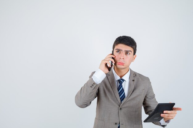 Chico joven en traje formal hablando por teléfono, sosteniendo la calculadora, pensando en algo