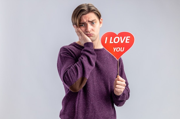 Foto gratuita chico joven triste en el día de san valentín con corazón rojo en un palo con texto te amo poniendo la mano en la mejilla aislado sobre fondo blanco.