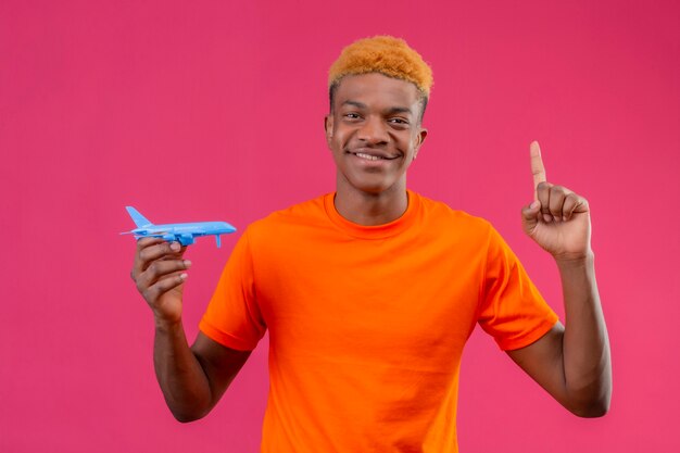 Chico joven viajero con camiseta naranja sosteniendo un avión de juguete apuntando con el dedo hacia arriba y sonriendo de pie sobre la pared rosa