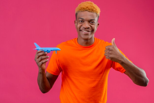 Chico joven viajero con camiseta naranja sosteniendo un avión de juguete sonriendo mostrando los pulgares para arriba sobre la pared rosa