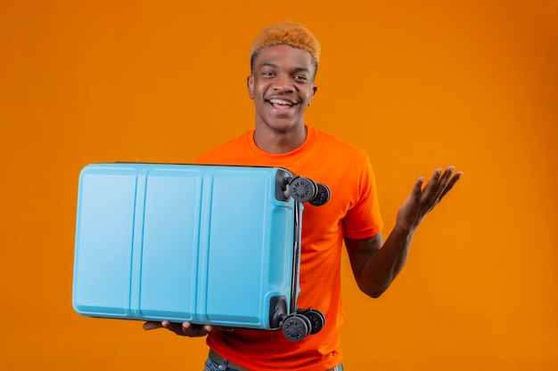 Chico joven viajero con camiseta naranja sosteniendo la maleta levantando la mano sonriendo alegremente de pie sobre la pared naranja