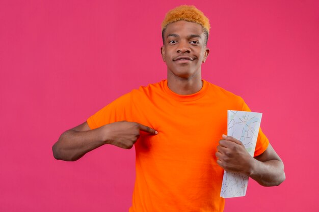 Chico joven viajero con camiseta naranja sosteniendo mapa sonriendo confiado de pie sobre la pared rosa