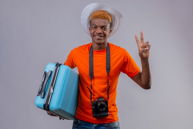Chico joven viajero guapo afroamericano en sombrero de verano con camiseta naranja con maleta de viaje mirando a cámara sonriendo amigable mostrando el número dos o el signo de la victoria sobre fondo blanco