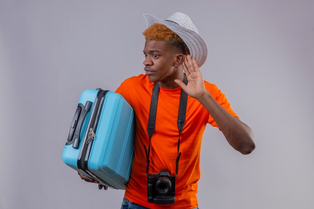 Chico joven viajero guapo afroamericano con sombrero de verano con camiseta naranja con maleta de viaje sosteniendo la mano cerca de su oreja tratando de escuchar la conversación de alguien sobre fondo blanco