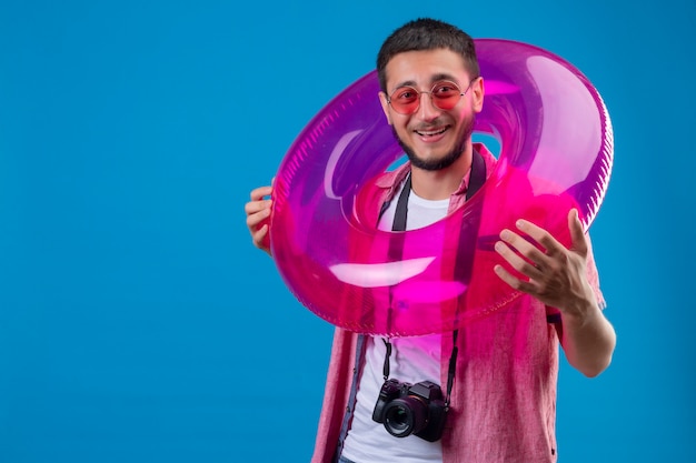 Chico joven viajero guapo con gafas de sol de pie con anillo inflable mirando a la cámara con cara feliz sonriendo alegremente de pie sobre fondo azul