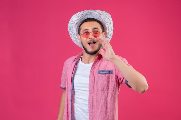 Chico joven viajero guapo con sombrero de verano con gafas de sol llamando a alguien con la mano cerca de la boca de pie sobre fondo rosa