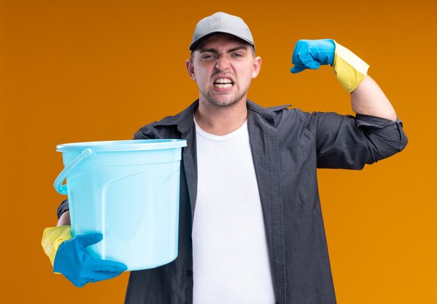 Chico de limpieza guapo joven confiado con camiseta y gorra con guantes sosteniendo un cubo que muestra un gesto fuerte aislado en la pared naranja