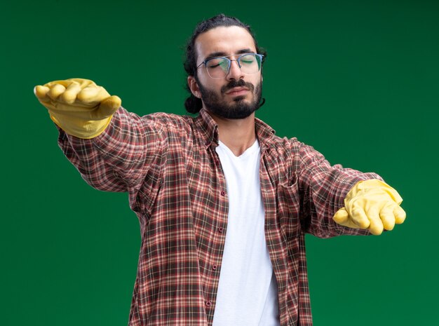 Chico de limpieza guapo joven durmiendo con camiseta y guantes tendiendo las manos aisladas en la pared verde