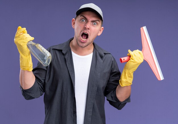 Chico de limpieza guapo joven enojado con camiseta con gorra y guantes sosteniendo la cabeza de la fregona y la botella de spray aislado en la pared púrpura