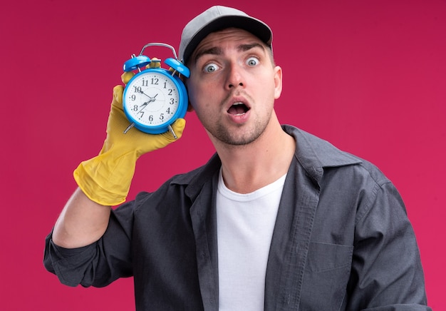 Foto gratuita chico de limpieza guapo joven sorprendido con camiseta y gorra con guantes sosteniendo y escuchando el despertador aislado en la pared rosa