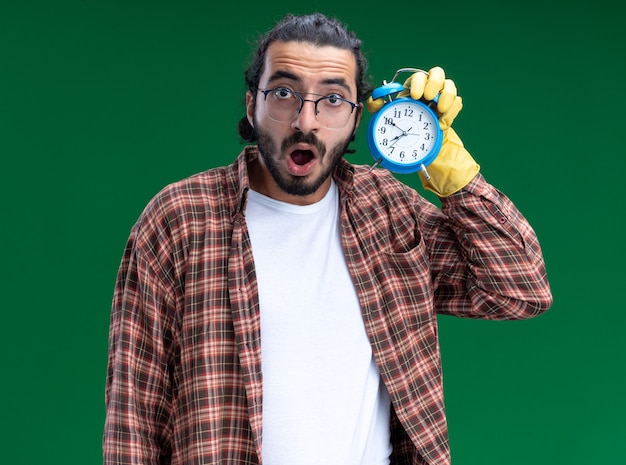 Chico de limpieza guapo joven sorprendido con camiseta y guantes sosteniendo y escuchando el despertador aislado en la pared verde