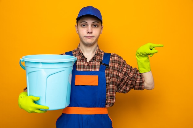 Chico de limpieza joven con uniforme y gorra con guantes sosteniendo balde aislado en la pared naranja con espacio de copia