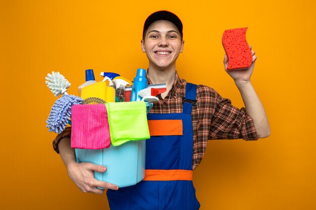 Chico de limpieza joven con uniforme y gorra sosteniendo un cubo de herramientas de limpieza con una esponja aislada en la pared naranja