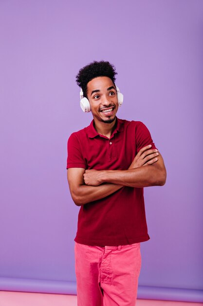 Chico lindo de buen humor posando con los brazos cruzados. Filmación en interiores de increíble modelo masculino escuchando música con sonrisa de sorpresa.