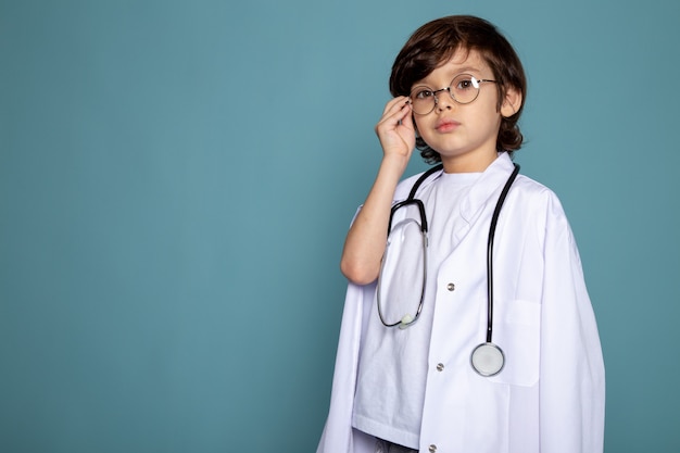 Chico lindo dulce niño adorable niño en traje médico blanco y gafas de sol en el escritorio azul