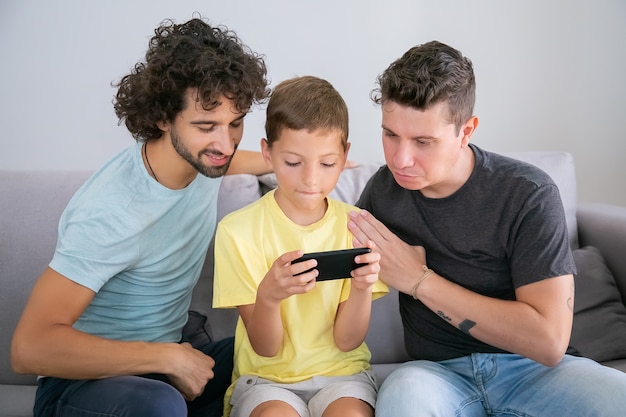 Chico lindo enfocado con smartphone, sus dos papás sentados a su lado y mirando la pantalla. Vista frontal. Concepto de familia y comunicación