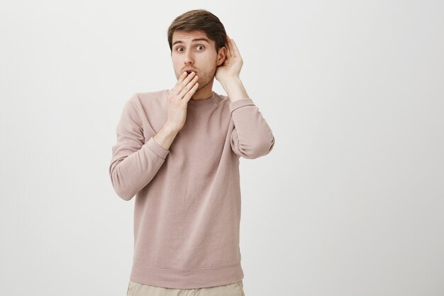 Chico lindo intrigado escuchando a escondidas, mantenga la mano cerca de la oreja