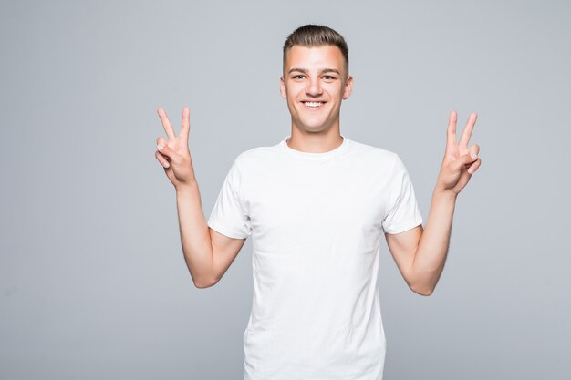 Chico lindo joven muestra el signo de la victoria aislado en la pared blanca