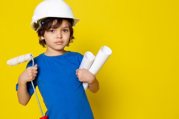 Chico lindo con pincel y planes de papel en casco blanco y camiseta azul sobre amarillo