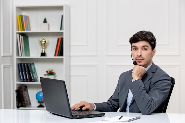 Chico lindo de servicio al cliente en traje gris con computadora y auriculares mirando seriamente