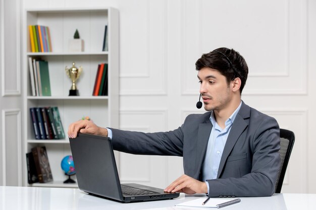 Chico lindo de servicio al cliente en traje gris con computadora y auriculares que terminan una videollamada