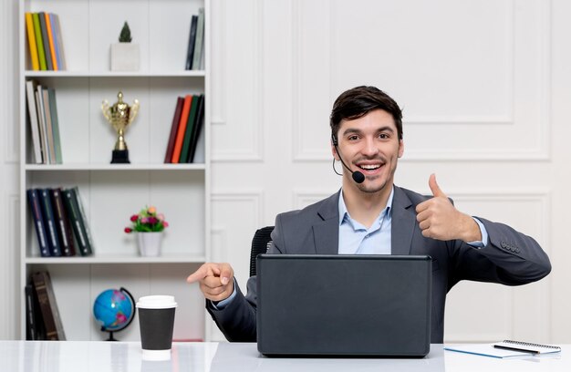 Chico lindo de servicio al cliente en traje gris con computadora y auriculares sonriendo y mostrando un buen gesto