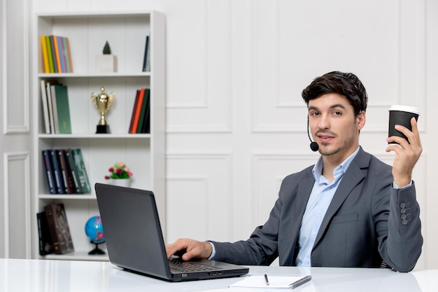 Chico lindo de servicio al cliente en traje gris con computadora y auriculares sosteniendo un vaso de papel